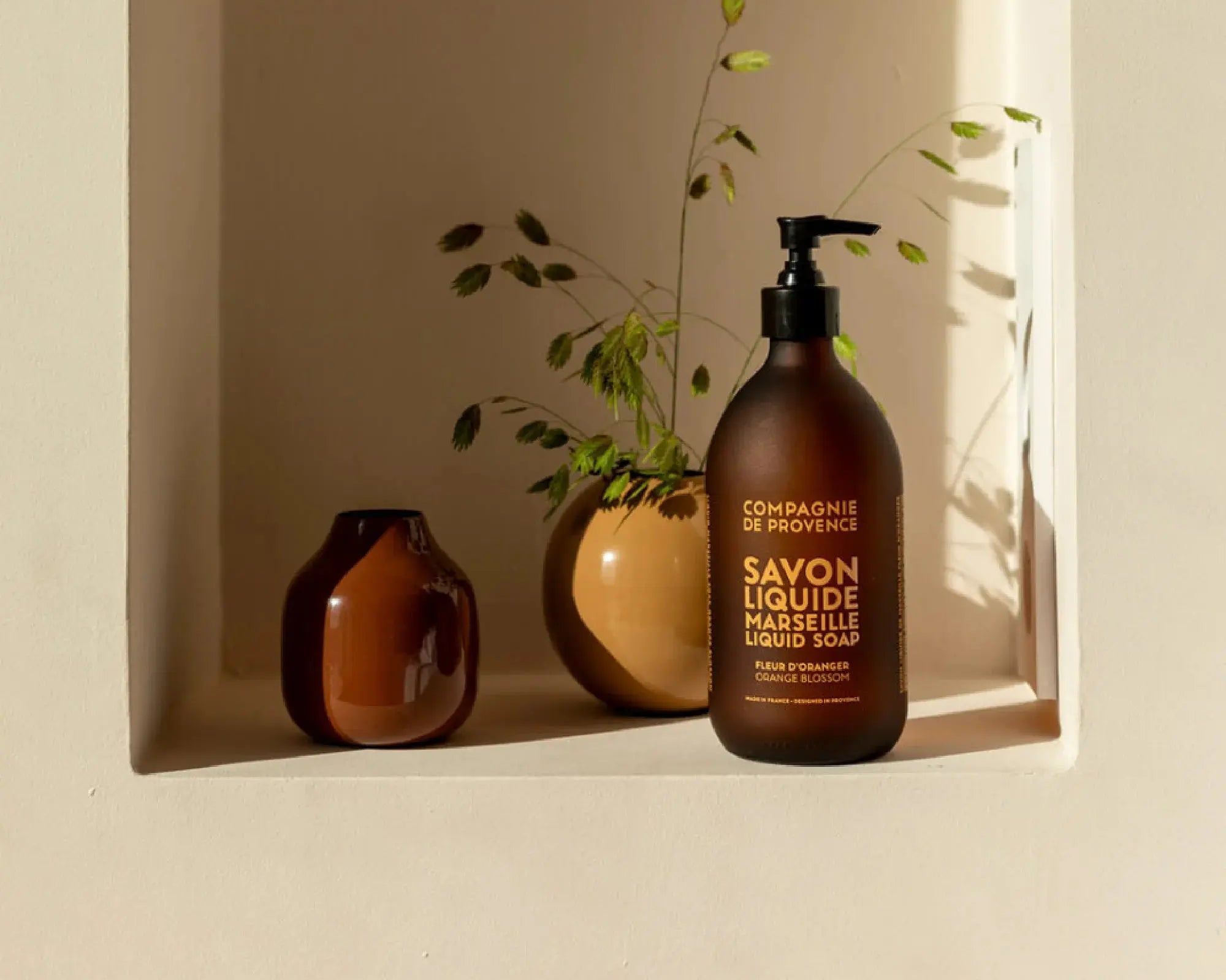 Savon liquide Fleur d'Oranger présenté dans une salle de bain lumineuse, posé près d'une plante et d'un autre flacon assorti.